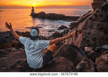 A Young Man Sitting On Some Rocks Watching The Orange Sunset Of The Pasajes San Juan Lighthouse, Nea