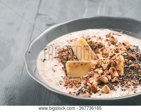 Close Up View Of Overnight Oats In Bowl With Banana, Lsa, Chia Seeds, Almond And Honey On Gray Woode