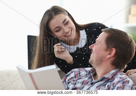 Guy Is Sitting On Couch With Book, Girl Is Smiling. Married Couple Having Fun At Home During Quarant