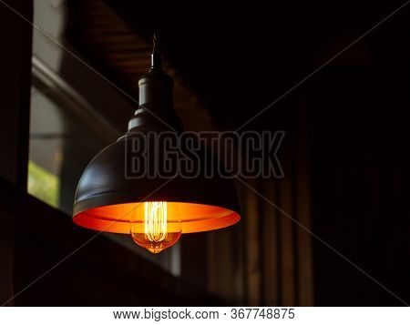 A Light Source In A Metal Lampshade Hangs From The Ceiling In A Dark Room. Yellow Light Bulb. Copisp