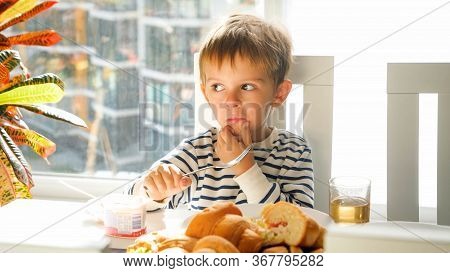 Portrait Of Little Thoughtfull Boy Sitting Behinddining Table And Having Breakfast