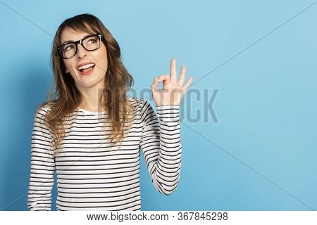Portrait Of A Young Friendly Woman With A Smile In A Casual T-shirt And Glasses Makes An Ok Gesture 