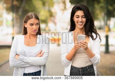 Envy Concept. Jealous Girl Looking At Friends Phone Walking Together Outdoors At Park. Selective Foc