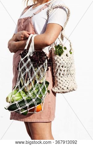 Young Woman In Light Summer Clothes Is Holding Mesh Shopping Bag With Vegetables, Greens Without Pla