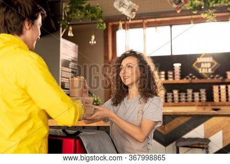 Young friendly waitress passing courier containers with ordered food and drinks while looking at him and helping with packing bag