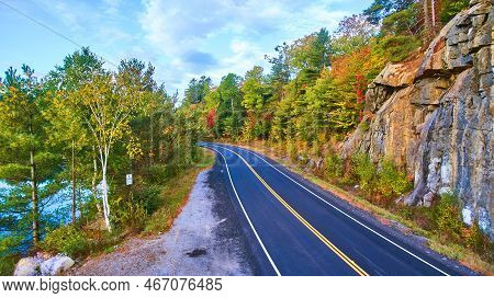 Drone Above Clean Road Going Through Fall Forest With Lake And Cliffs Around