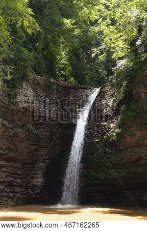 Landscape With Summer Hight Waterfall In Lush Green Forest With Sunbeams, Splashes And Layered Rocks