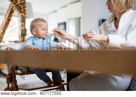 Mother Wearing Cosy Bathrope Spoon Feeding Her Baby Boy Child In Baby High Chair With Fruit Puree At
