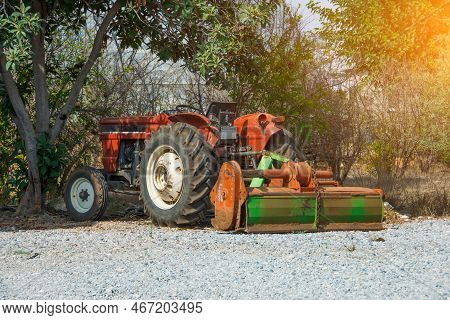Red Old Tractor With A Plow Is Parked Near The Trees And Bushes In The Village.