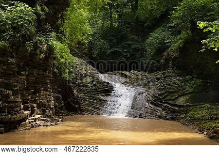 Landscape With Summer Waterfall In Lush Green Forest With Sunbeams, Splashes And Layered Rocks Overg