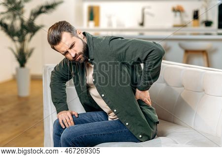 Back Problems Concept. Sad Caucasian Man, In Casual Wear, Sit On Sofa In Living Room, Experiencing A