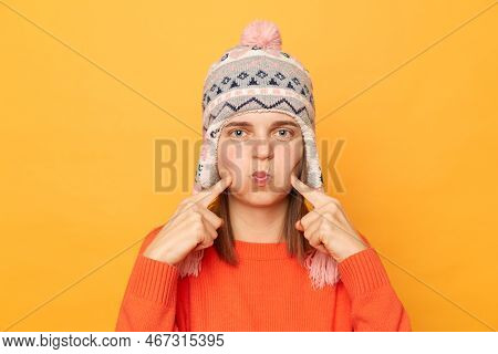 Indoor Shot Of Funny Woman Wearing Orange Sweater And Knitted Earflap Hat Standing Isolated Over Yel