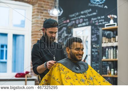 Professional Barber Preparing His Happy Indian Client In Yellow Cape For A Hearcut In A Barbershop. 