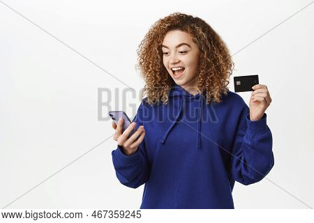 Portrait Of Curly Girl Smiles, Makes Order, Shops Online In App, Shows Credit Card And Holds Mobile 