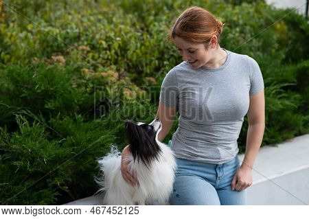 Caucasian Red-haired Woman Cuddling With Pappilion Dog Outdoors. Black And White Continental Spaniel