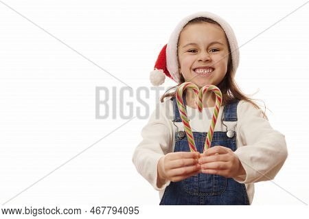 Mischievous Little Child Girl Wearing Santa Hat, Holding Red And Green Strippes Candy Canes In Shape