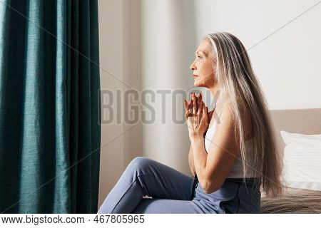 Side View Of A Senior Woman With Long Hair Looking Thoughtful While Sitting On A Bed
