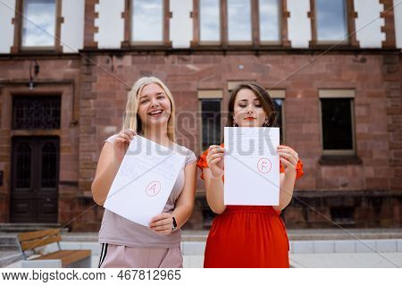 Two Students Near University With Test Results. One Woman Happy Because Of High Mark And The Other I