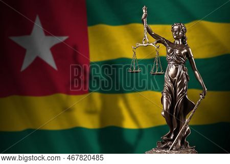 Togo Flag With Statue Of Lady Justice And Judicial Scales In Dark Room. Concept Of Judgement And Pun