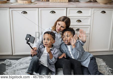Happy Multinational Family Of Mother And Two Sons Sitting On Warm Floor In Light Kitchen During Week