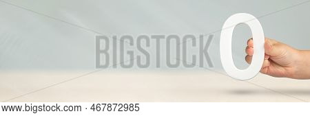 Numeral Zero In Hand. A Hand Holds A White Number Zero On A Blurred Background With Copy Space. Zero