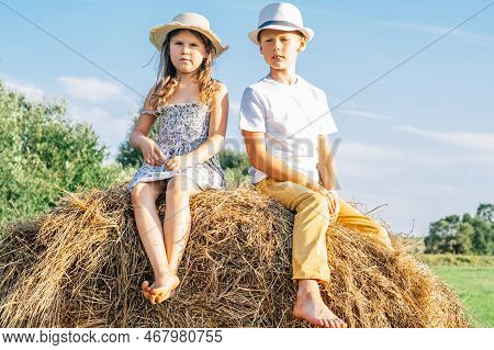 Cute Little Girl And Boy Sitting Together At Top Of Haystack. Friends Resting On Hayrick. Outdoor Wa
