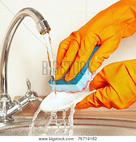 Hands in rubber gloves with sponge wash dirty dishes under running water
