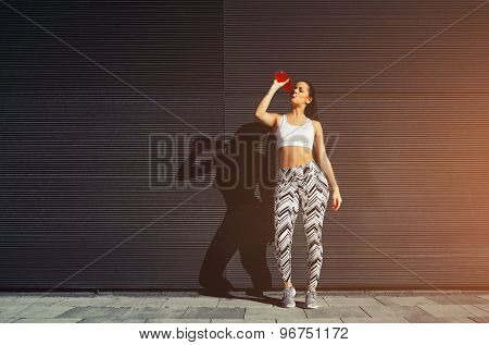 Young fit woman with beautiful figure refreshing with energy drink against black wall in the city