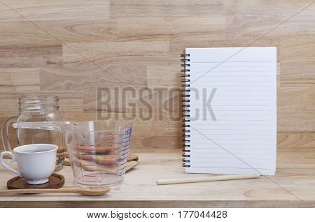 blank notebook with coffice cup and measuring cup and wooden spoon cooking supplies on wood background.