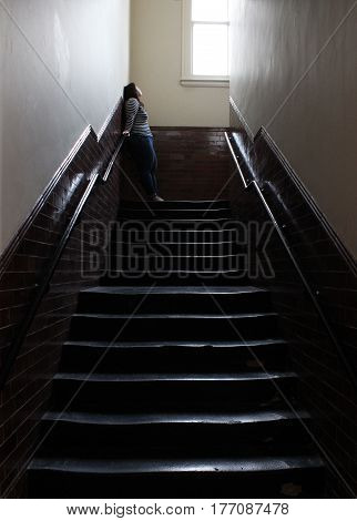 Brunette woman at the top of a dark staircase in an old unused school looks up at a high window