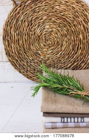 Stack of folded linen towels wicker rattan coaster on white wood background twig of rosemary kitchen interior concept