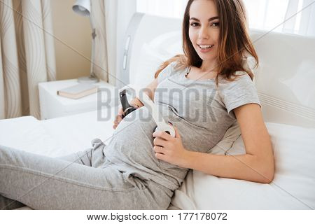 Smiling pregnant woman holding headphones on her tummy and looking at camera while lying on bed