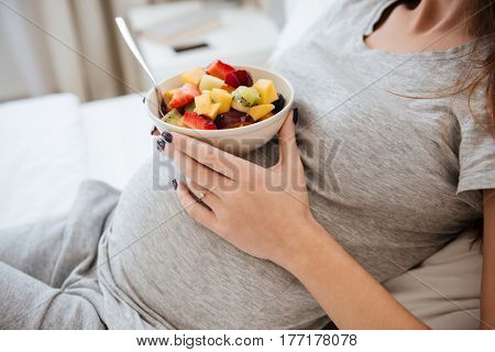 Side view of pregnant woman which holding a plate with fruits on her tummy. Cropped image
