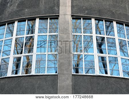Fragment of roundish gray wall of old building with big windows overlooking the park. Glasses in white wooden frames reflect blue sky and trees. Image can be used as a background.