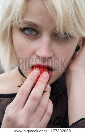 Beautiful young Russian girl with blond hair and blue eyes eating a strawberry in the field. She is dressed in a transparent black dress.