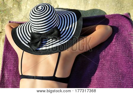 Young attractive woman in a large black and white striped summer hat wearing a black bikini is sunbathing on the beach.