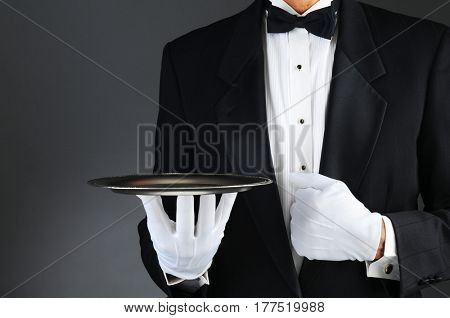 Closeup of a tuxedo wearing waiter holding a silver tray in front of his body. Horizontal format on a light to dark gray background.