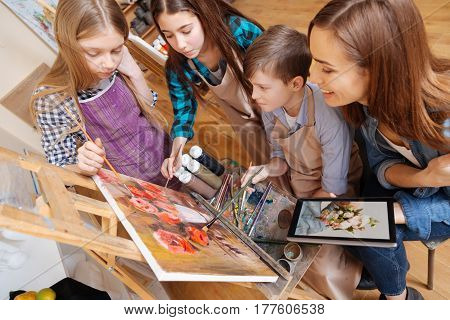 Enjoying art lesson . Positive helpful attentive teacher sitting in the art school and expressing joy while teaching painting kids and using gadget
