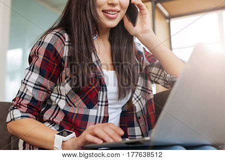 In the office. Happy pretty delighted woman having a phone conversation and using the laptop while working