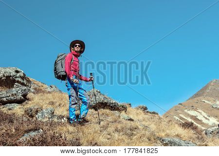 Young Hiker in red Jacket and Blue trekking Pants carrying Backpack and walking Sticks staying on grassy Slope