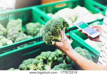 Young woman shopping healthy food in supermarket blur background. Female hands buy products broccoli using smartphone in store. Hipster at grocery using smartphone. Person comparing price of produce