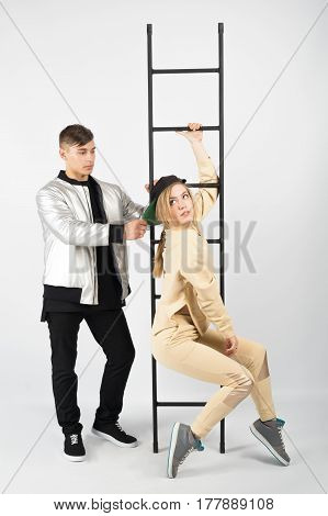 Modern fashionable hipster couple with ladder. Young muscular man wearing black clothes and sneakers with girl in yellow clothes on white background. He is holding her snapback