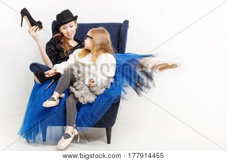 The elder sister and little girl ballerina are sitting in a chair. They keep their shoes on high heels. Girls in ballet tutu and pointe shoes. The sisters are talking
