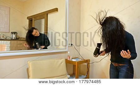Beautiful brunette woman drying long black silky hair with hairdryer and smartens up in front of mirror in make-up dressing room.