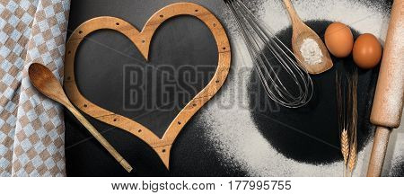 Baking background with empty blackboard in the shape of a heart on a black table with rolling pin whisk eggs spoons ears of wheat and a checkered tablecloth
