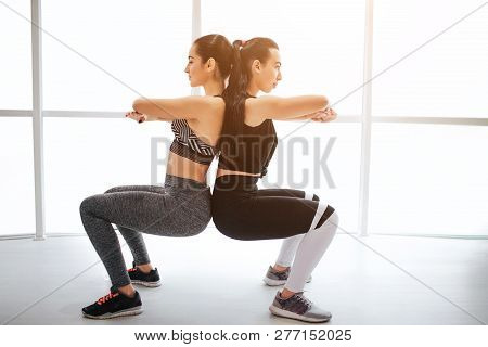 Two Sloim Young Women Exerising In Fitness Room. They Keep Hands Crossed In Chest Area And Touch Eac