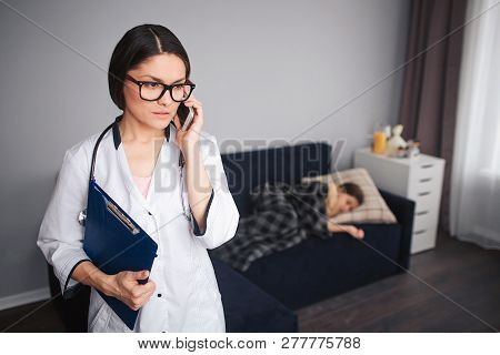 Serius Worried Young Woman Talk On Phone. She Stand In Room. Her Patient Lying On Couch Behind. Smal