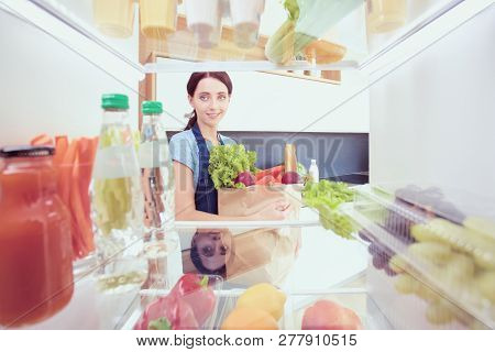Portrait Of Female Standing Near Open Fridge Full Of Healthy Food, Vegetables And Fruits. Portrait O