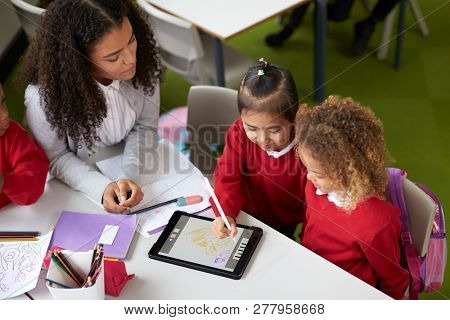 Elevated view of two girls using a tablet computer sitting at table in an infant school class a with female teacher helping them