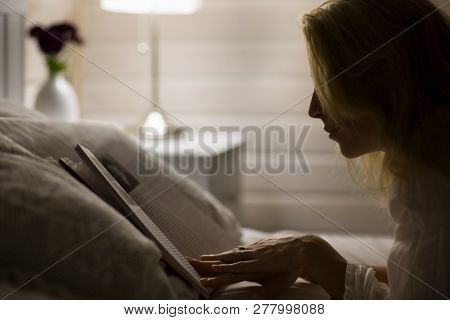 Beautiful Long Haired Blonde Woman Lays On Bed, And Read Book In Dark Room At Night.